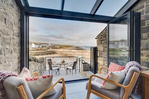 Une terrasse fermée par des baies vitrées, avec une table et des chaises, offrant une vue sur la plage et le paysage côtier.