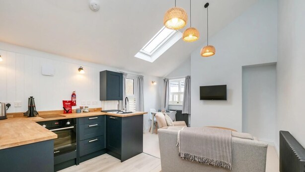 A modern kitchen with dark cabinets, a wooden countertop, and a built-in oven. There is a grey sofa with a striped throw blanket and a flat-screen TV mounted on the wall. - 1 Bed in Sheringham (oc-n33618) (Sheringham)