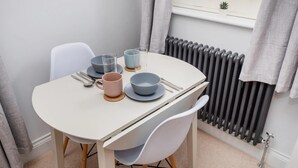 A dining table with plates, bowls, and cutlery, a chair, and a radiator in a room with a window and curtains. - 1 Bed in Sheringham (oc-n33618) (Sheringham)