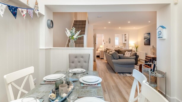 Un comedor con una mesa dispuesta para cuatro personas, un sofá tapizado gris al fondo, y una escalera que sube a un nivel superior.