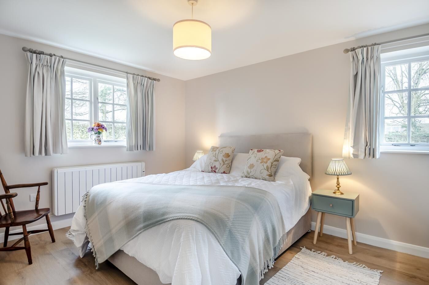 Ein Schlafzimmer mit einem großen Bett, einem Nachttisch mit Lampe, einem Sessel und einem Fenster mit Vorhängen.