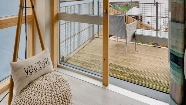 A room with large windows, a wooden floor, a round pouf, a lamp, and a view of the balcony and outside. - 2 Bed in Seahouses (oc-566411) (Seahouses)
