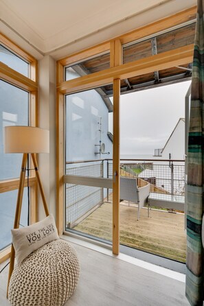 Ein Zimmer mit großen Fenstern, Holzboden, einem runden Pouf, einer Lampe und Blick auf Balkon und Außenbereich.