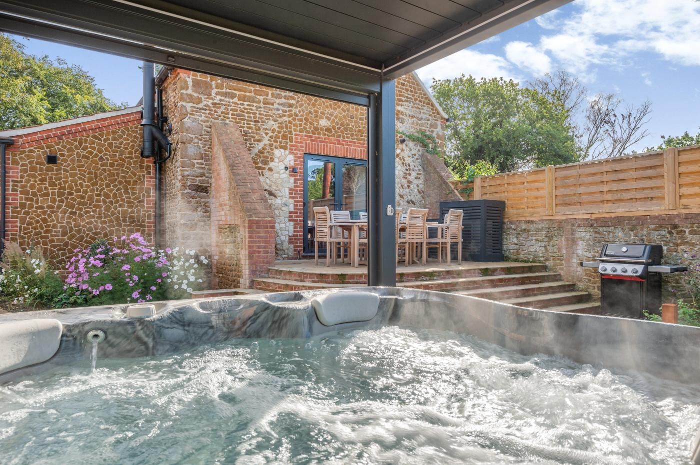 A hot tub with a stone wall and wooden deck, outdoor seating, and a barbecue grill.