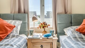 Una habitación con dos camas, una mesita de noche con libros y una lámpara, y una ventana con cortinas.