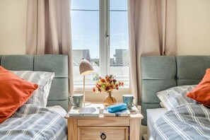 A room with two beds, a nightstand with books and a lamp, and a window with curtains.