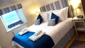 Een net opgemaakt bed met blauw-witte beddengoed, een raam met uitzicht op zee en een nachtkastje met een lamp en boeken.