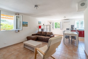 Ein Wohnzimmer mit Sofa, Couchtisch, Esstisch mit Stühlen, einer Küchenzeile und einem großen Fenster mit Blick ins Grüne.