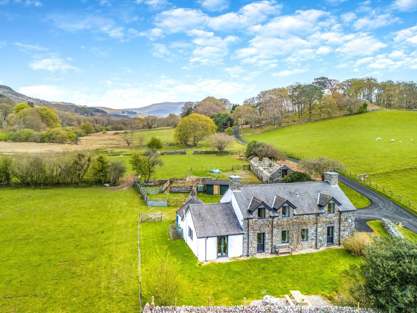 A stone house with a sloped roof, surrounded by green fields and a winding road.