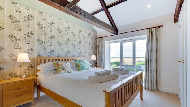 A bedroom with a wooden bed, two bedside tables with lamps, a large window with curtains, and a view of the countryside. - 4 Bed in nr Beadnell (oc-566760) (Beadnell)