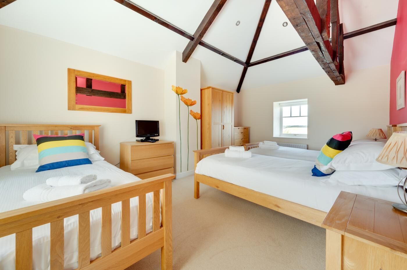 Une chambre avec deux lits en bois, un téléviseur posé sur une table de chevet en bois, une armoire et une fenêtre laissant entrer la lumière naturelle.