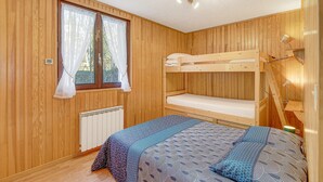 Quarto com paredes de madeira, uma cama beliche, janela com cortinas, radiador e abajur.