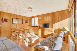 A wooden-paneled room with a sofa, a dining table with chairs, a TV, and a window. - Apartment 'Orcières Village' with Mountain View and Balcony (Orcières)