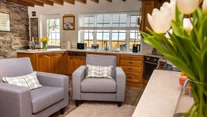 A cozy living room with two grey armchairs, a wooden cabinet, and a kitchen area with a window. - 3 Bed in Cwmystradllyn (oc-wag708) (Cwmystradllyn)