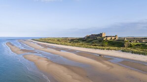 A coastal landscape with a sandy beach, a castle-like structure, and a clear sky. - 2 Bed in Bamburgh (oc-nb1969) (Bamburgh)