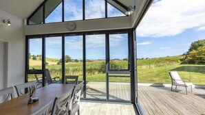 A spacious room with a large table and chairs, a clock on the wall, and a view of a green landscape through large windows. - 2 Bed in Bamburgh (oc-nb1969) (Bamburgh)
