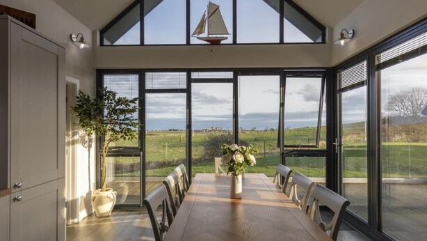 A dining area with a long table, chairs, and a vase of flowers. There are large windows with a view of a green landscape. - 2 Bed in Bamburgh (oc-nb1969) (Bamburgh)