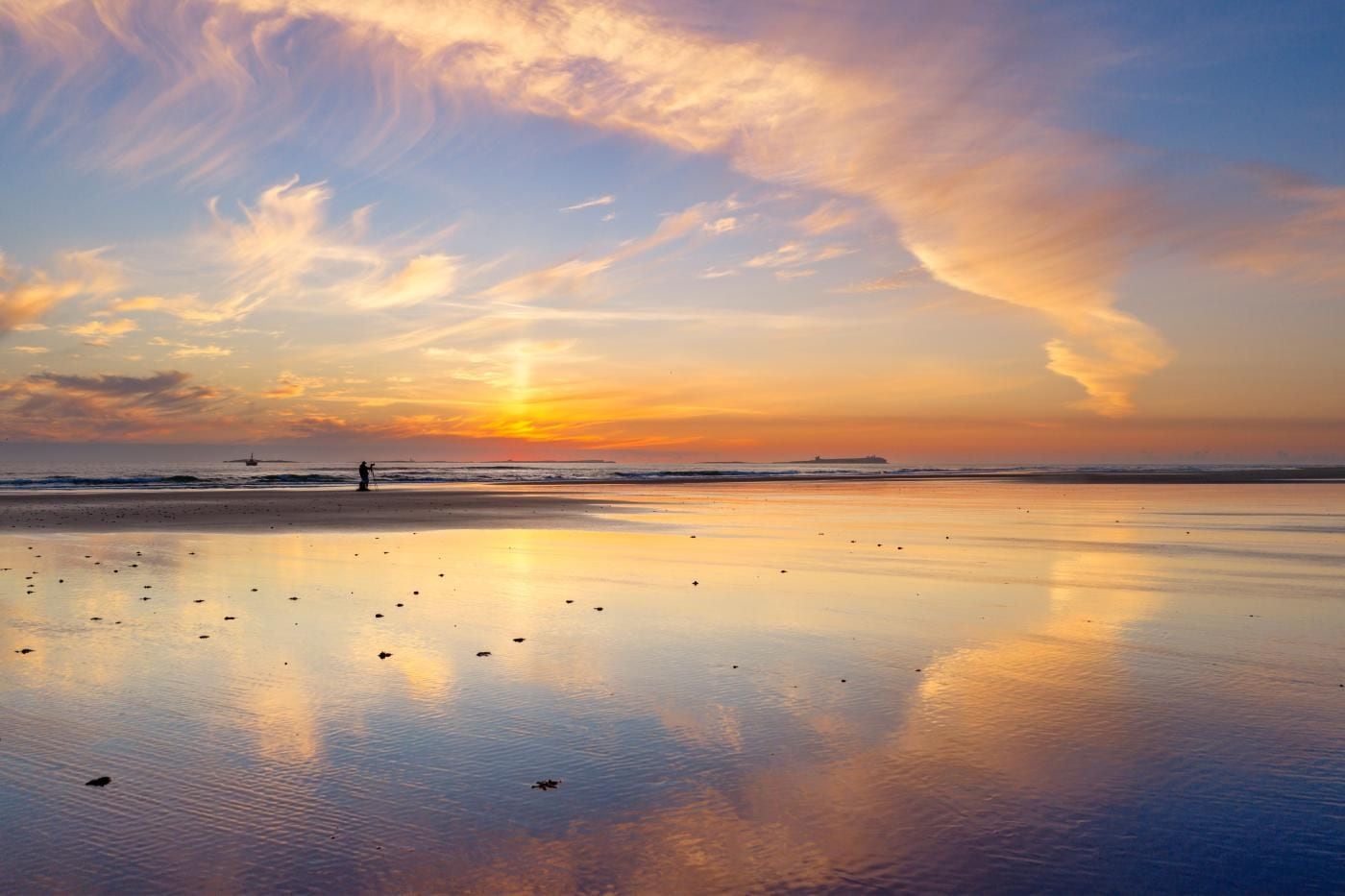 A beach with calm waters reflecting the sky, a person walking, and a distant ship.