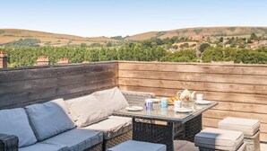 A rooftop patio with a wicker sofa, a glass table, and a view of rolling hills and buildings. - 2 Bed in Swanage (oc-wy619) (Swanage)