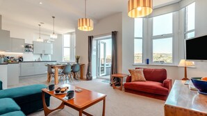 A modern living room with a red sofa, teal armchair, and wooden coffee table. There is a kitchen area with white cabinets and a dining table. Large windows provide a view of the outdoors. - 2 Bed in Swanage (oc-wy619) (Swanage)