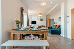 A modern dining area with a wooden table set for four, a bench, and a TV in the background. - 2 Bed in Swanage (oc-wy619) (Swanage)