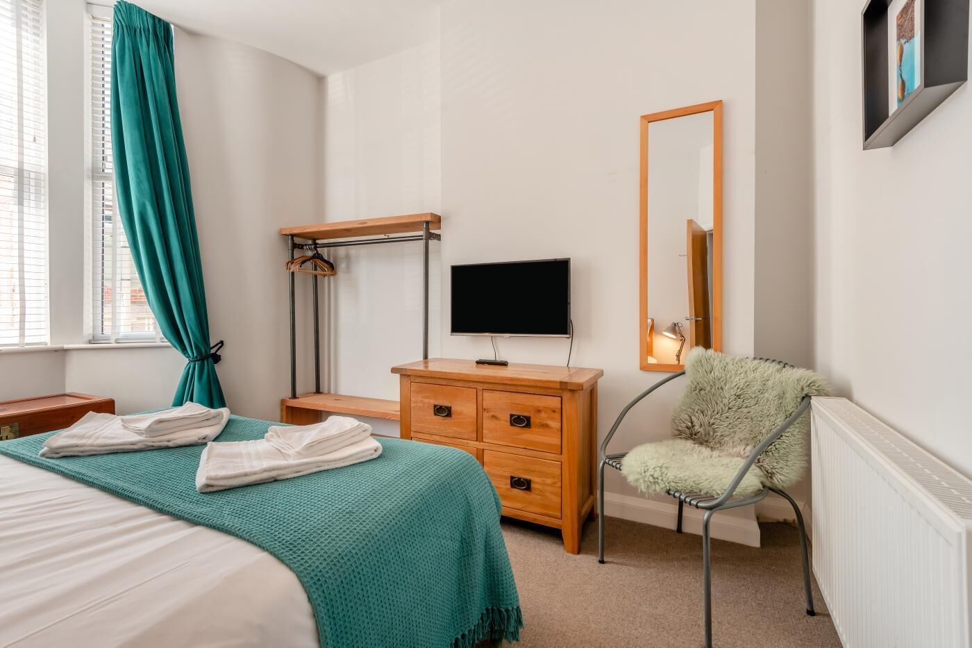 A bedroom with a bed, a wooden dresser, a television, a mirror, and a fluffy chair.