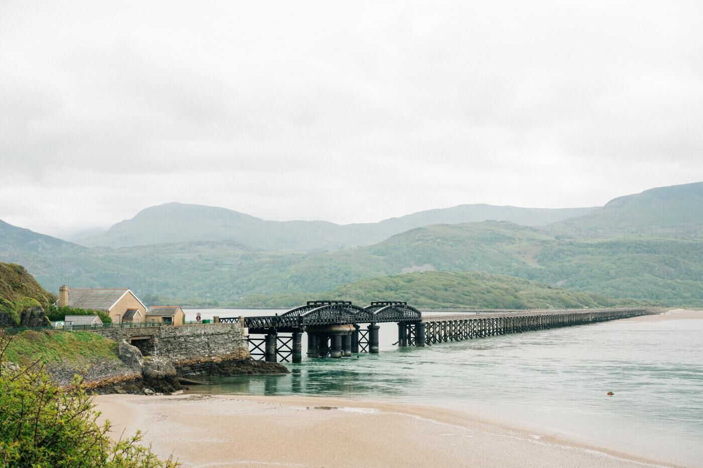 Un paisaje costero con un puente de piedra sobre el agua, una playa y una casa con muro de piedra.