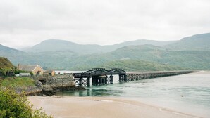 Un paisaje costero con un puente de piedra sobre el agua, una playa y una casa con muro de piedra.