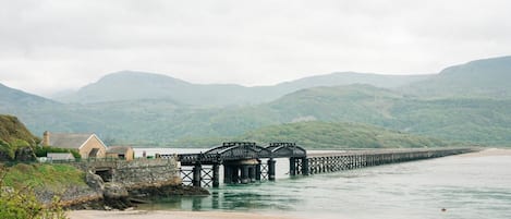 A coastal scene with a stone bridge over water, a beach, and a house with a stone wall.
