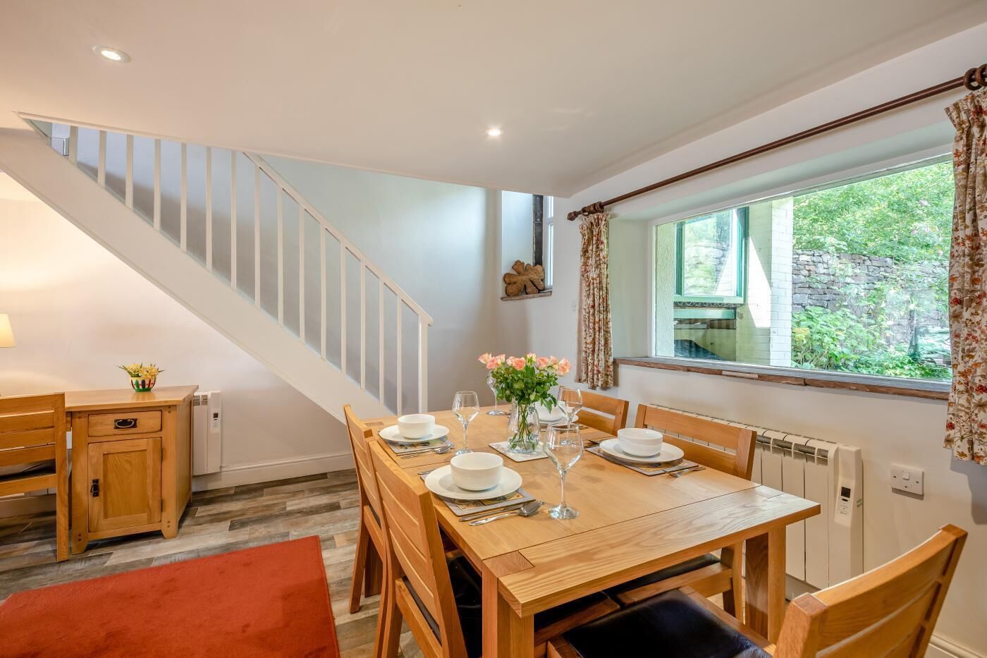 A dining area with a wooden table set for four, a staircase, and a window with curtains.