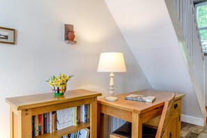 Ein Eckzimmer mit einem Holztisch, einem Bücherregal, einer Lampe und einer Vase mit Blumen.