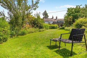 A grassy outdoor area with a chair and a table, surrounded by lush greenery and a house in the background. - 2 Bed in Pooley Bridge (oc-r33740) (Pooley Bridge)