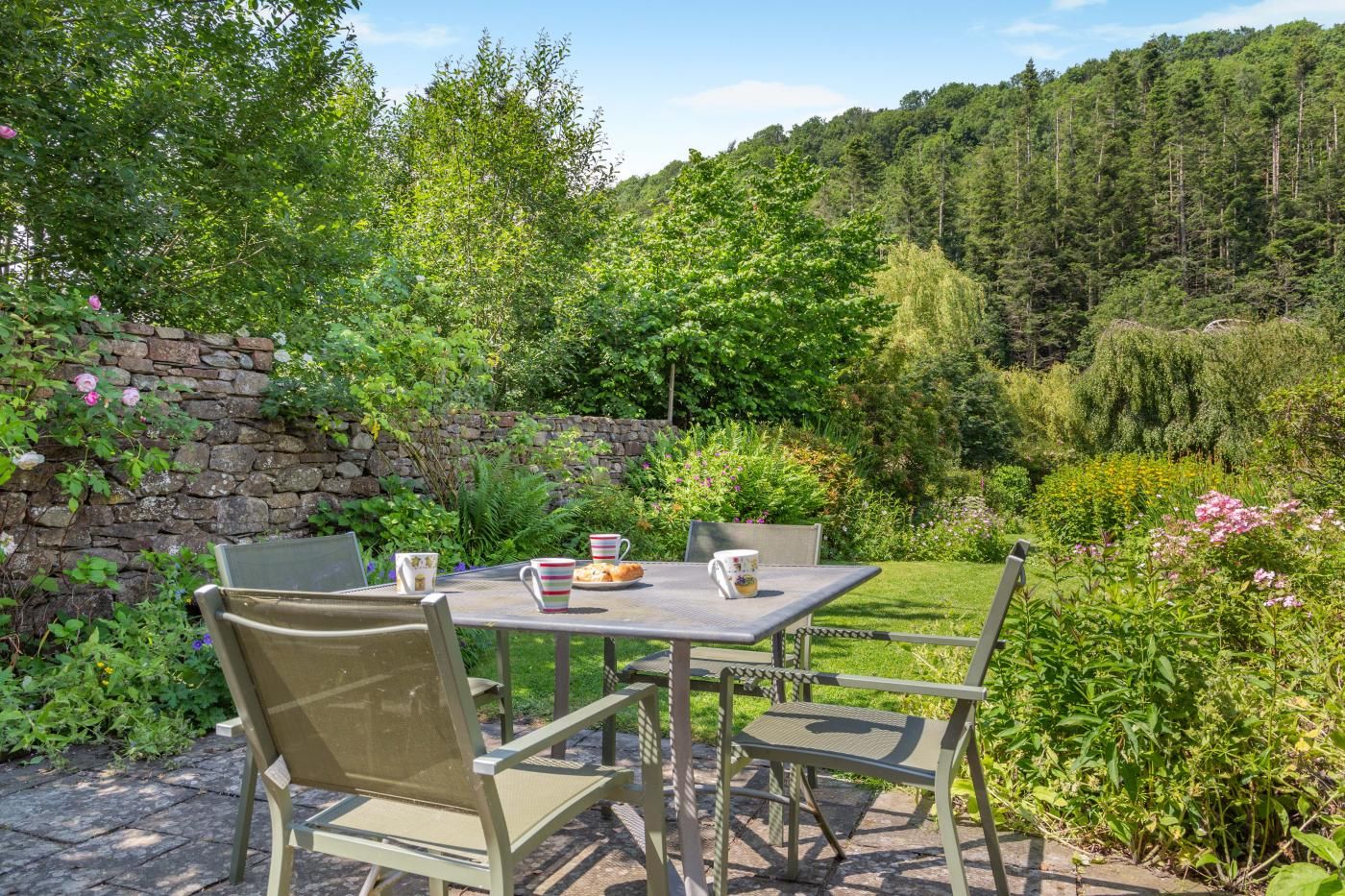 An outdoor seating area with a table and chairs, surrounded by lush greenery and a stone wall.