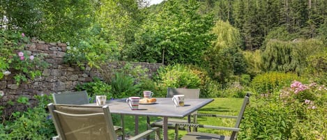 An outdoor seating area with a table and chairs, surrounded by lush greenery and a stone wall.