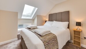 Ein modernes Schlafzimmer mit Dachfenster, einem großen Bett mit weißen Bettwäsche, einem Nachttisch mit Lampe und Blick zum Himmel durch das Dachfenster.