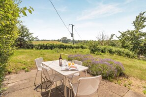 An outdoor patio with a table set for two, surrounded by lush greenery and purple flowers.