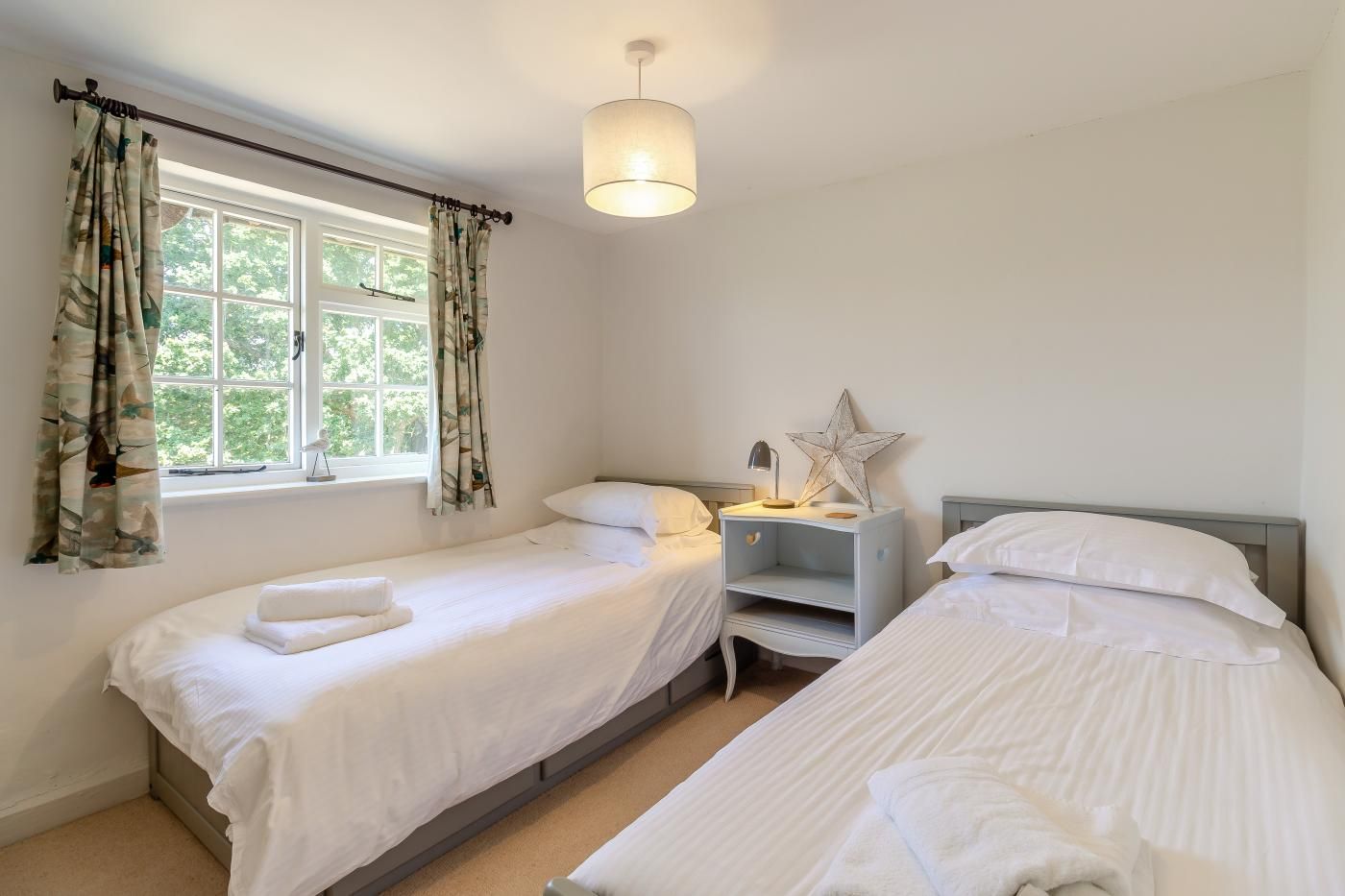 Une chambre avec deux lits simples, une table de chevet avec une lampe et une fenêtre avec des rideaux.