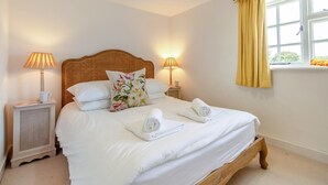 Un llit ben fet amb llençols blancs, dues làmpades de nit, un coixí floral i una finestra amb cortines grogues.