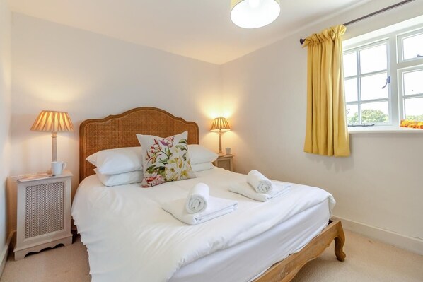 A neatly made bed with white linens, two bedside lamps, a floral pillow, and a window with yellow curtains.