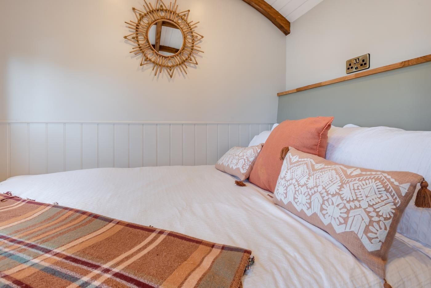 A bedroom with a bed, pillows, a blanket, a wall-mounted mirror, and a power outlet.