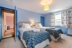 A bedroom with two beds, a door leading to another room, a window with floral curtains, and a ceiling light. - 3 Bed in Weymouth (oc-s33545) (Weymouth)