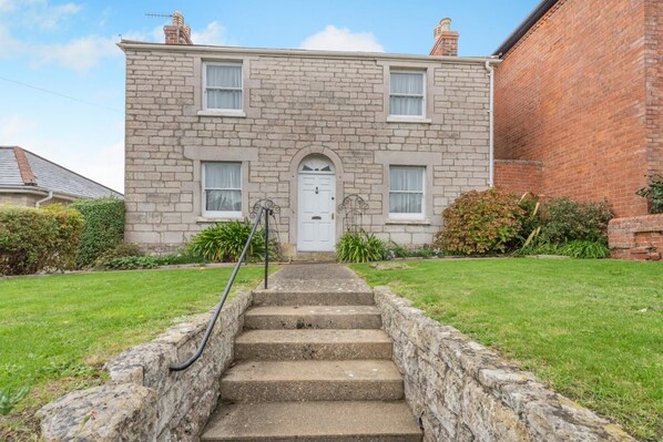 A two-story stone house with a white door and windows, a front garden, and a brick wall to the right. - 3 Bed in Weymouth (oc-s33545) (Weymouth)