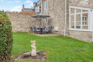 A stone building with a stone wall, a grassy yard, a stone statue, and a table with chairs and an umbrella. - 3 Bed in Weymouth (oc-s33545) (Weymouth)