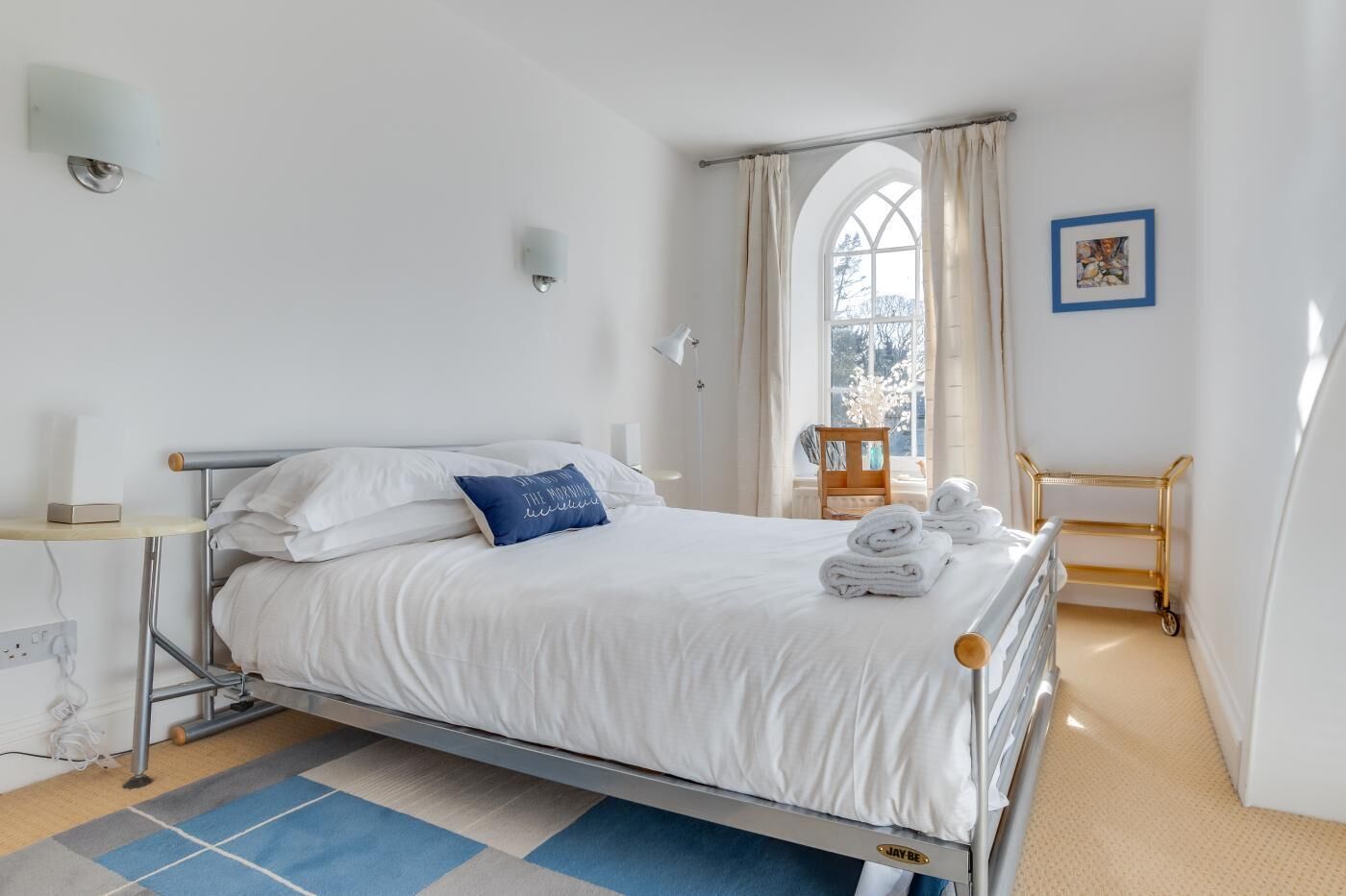 A neatly made bed with white linens and a blue pillow, a small bedside table with a lamp, a window with curtains, and a framed picture on the wall.