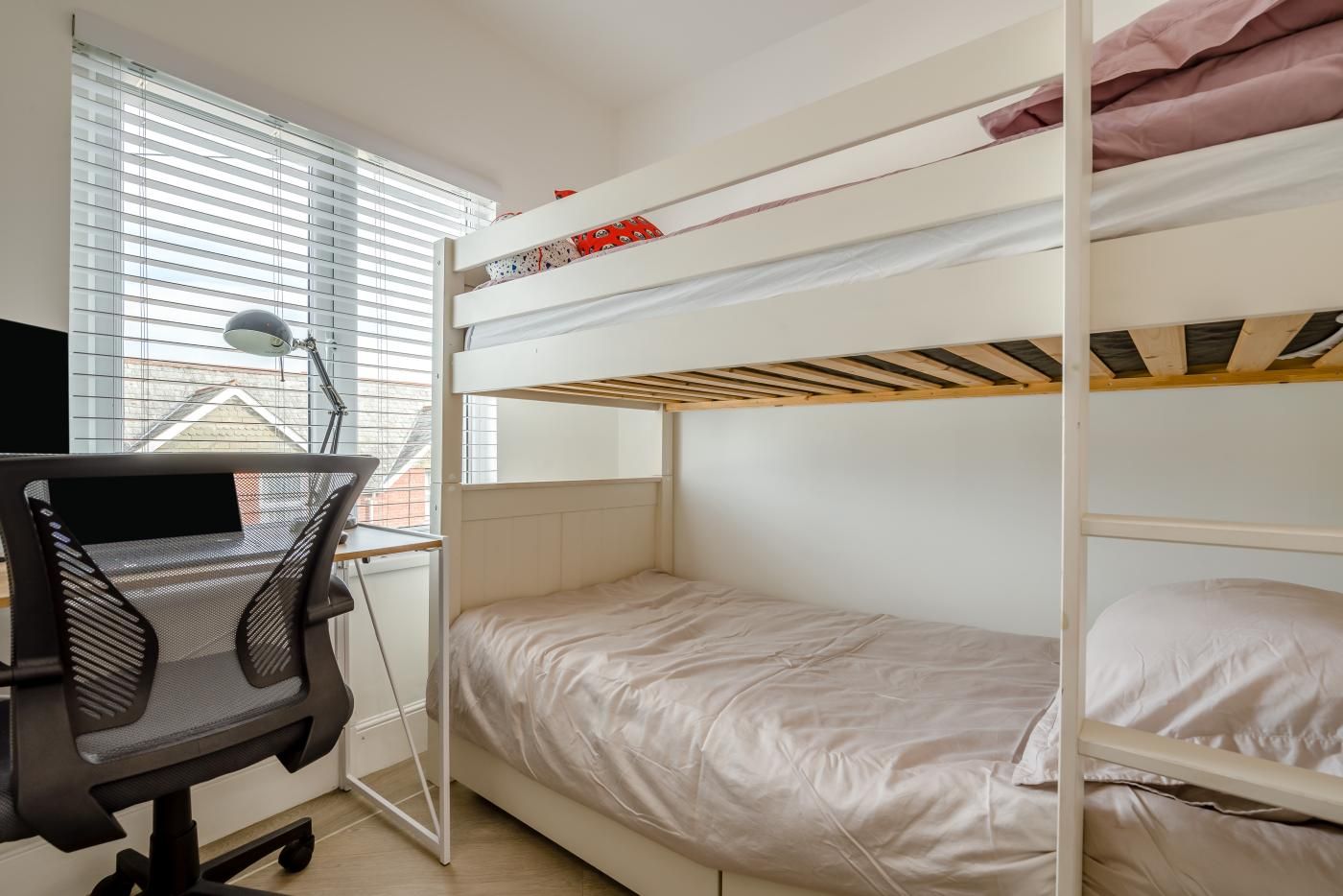 A bunk bed room with a desk and chair, a window with blinds, and a lamp.