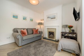 A living room with a grey sofa, a fireplace, a flat-screen TV mounted on the wall, and a small table with a vase of flowers. - 3 Bed in Padstow (oc-f33036) (Padstow)