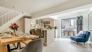 A modern kitchen with white cabinets, a dining area with a wooden table set for four, and a blue armchair in a bright living space with a staircase. - 3 Bed in Padstow (oc-f33036) (Padstow)