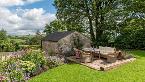 A stone building with a patio area, including a round table and wicker chairs, surrounded by a well-maintained garden with various flowers and a large tree.