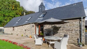 Un bâtiment en pierre doté d’un toit en pente, d’une cheminée et d’un patio aménagé avec des meubles d’extérieur et des plantes en pot.