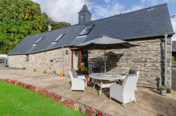 A stone building with a sloped roof, a chimney, and a patio area with outdoor seating and potted plants. - 2 Bed in Machynlleth (oc-491245) (Llanwrin)
